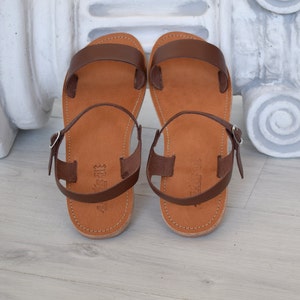 K&ouml;nnte beinhalten: Ein Paar braune Ledersandalen mit hellbraunem Fu&szlig;bett. Die Sandalen haben ein schlichtes Design mit einem Riemen &uuml;ber den Zehen und einem Kn&ouml;chelriemen mit einer silbernen Schnalle. Die Sandalen werden auf einer hellen Holzoberfl&auml;che pr&auml;sentiert.