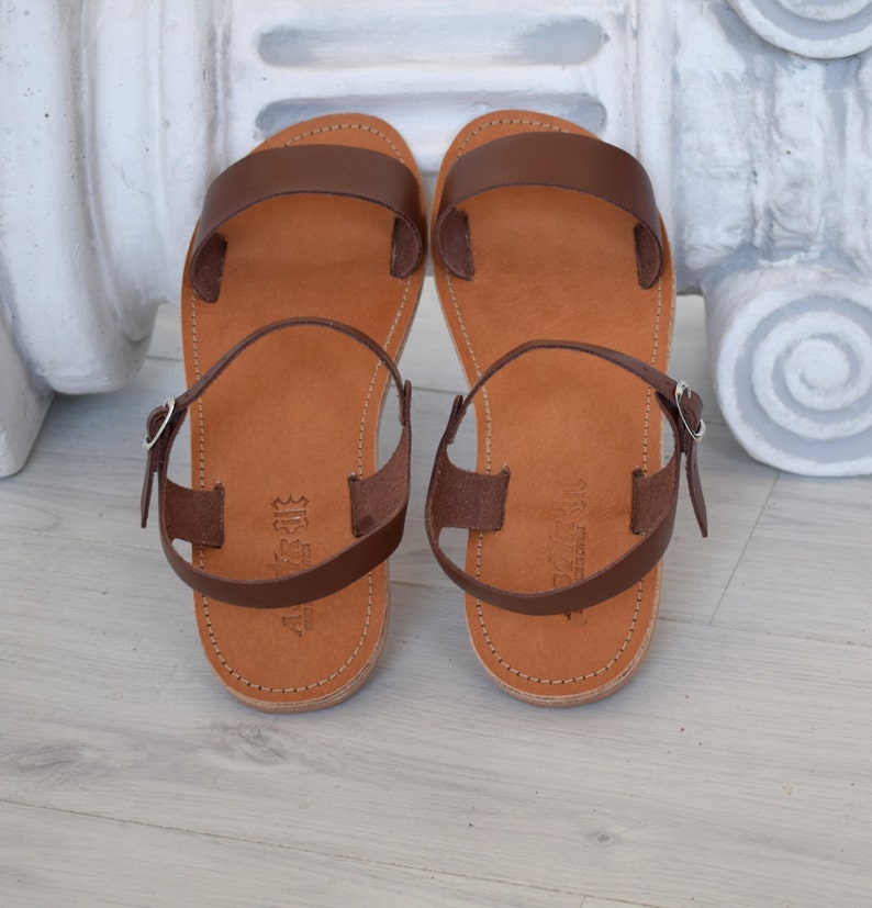 K&ouml;nnte beinhalten: Ein Paar braune Ledersandalen mit hellbraunem Fu&szlig;bett. Die Sandalen haben ein schlichtes Design mit einem Riemen &uuml;ber den Zehen und einem Kn&ouml;chelriemen mit einer silbernen Schnalle. Die Sandalen werden auf einer hellen Holzoberfl&auml;che pr&auml;sentiert.