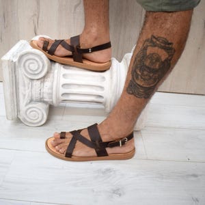 May include: Dark brown leather sandals featuring a toe loop design, multiple crisscrossing straps across the foot, and an adjustable ankle strap with a silver-toned metal buckle. The sandals have a flat, light tan sole and a minimalist construction.