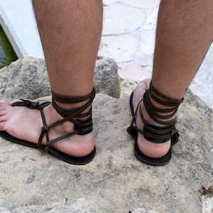 Puede incluir: Sandalias de cuero marr&oacute;n oscuro con correas envolventes en el tobillo. Las sandalias tienen un dise&ntilde;o sencillo con suela plana y una correa para el dedo del pie tipo tanga. Las correas se envuelven alrededor de los tobillos y las pantorrillas.