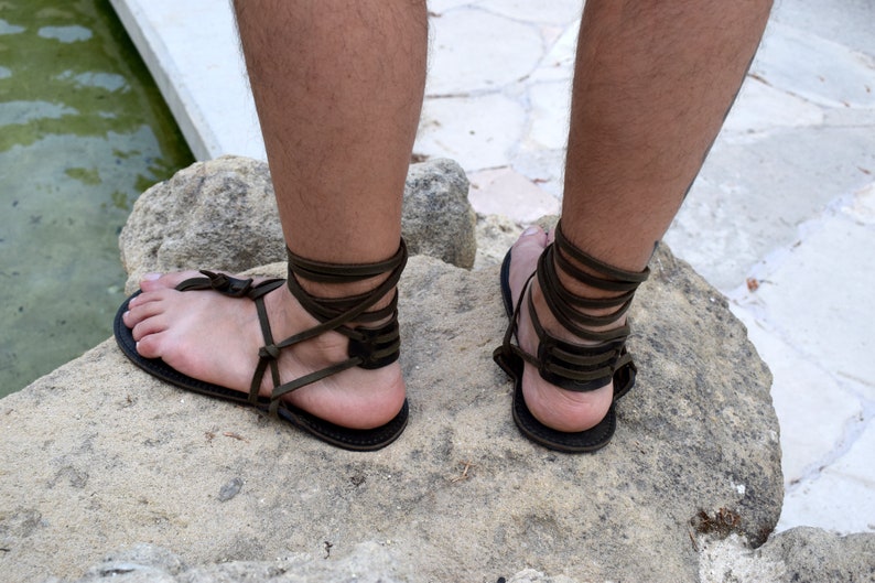 Puede incluir: Sandalias de cuero marr&oacute;n oscuro con correas envolventes en el tobillo. Las sandalias tienen un dise&ntilde;o sencillo con suela plana y una correa para el dedo del pie tipo tanga. Las correas se envuelven alrededor de los tobillos y las pantorrillas.