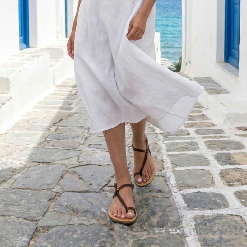 Puede incluir: Un vestido blanco de largo midi con una silueta holgada y fluida, combinado con sandalias de dedo de cuero marr&oacute;n oscuro que presentan tiras finas que rodean el pie y se aseguran con una peque&ntilde;a hebilla met&aacute;lica en el tobillo.