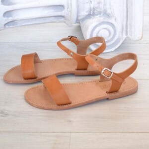 K&ouml;nnte beinhalten: Ein Paar hellbraune Ledersandalen mit schlichtem Design. Jede Sandale hat eine flache Sohle, einen Riemen &uuml;ber den Zehen und einen Kn&ouml;chelriemen mit silberner Schnalle. Die Sandalen werden auf einer hellen Holzoberfl&auml;che pr&auml;sentiert, ca. 24 cm lang.