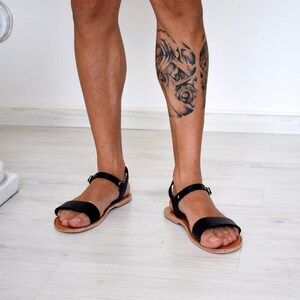 K&ouml;nnte beinhalten: Schwarze Ledersandalen mit schlichtem Design. Die Sandalen haben einen einzelnen Riemen &uuml;ber dem Fu&szlig; und einen verstellbaren Kn&ouml;chelriemen. Die Sohlen sind hellbraun. Die Sandalen werden an den F&uuml;&szlig;en einer Person gezeigt.