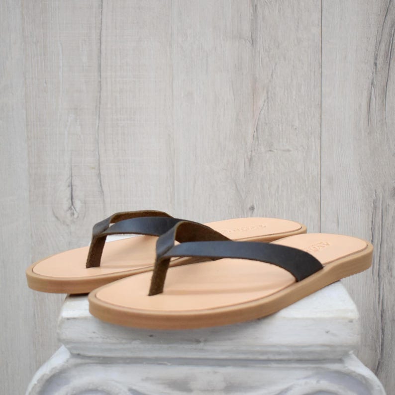 May include: A pair of leather flip-flops with dark brown straps and light brown soles. The sandals are displayed on a white pedestal against a light wood-grain background. The flip-flops are a neutral colour.