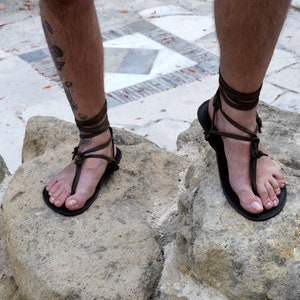 Puede incluir: Sandalias de cuero negro con correas envolventes verde oliva. Las sandalias tienen un dise&ntilde;o tipo tanga y se usan sobre una superficie de piedra. Las correas se envuelven alrededor de los tobillos.