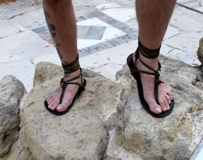 Puede incluir: Sandalias de cuero negro con correas envolventes verde oliva. Las sandalias tienen un dise&ntilde;o tipo tanga y se usan sobre una superficie de piedra. Las correas se envuelven alrededor de los tobillos.