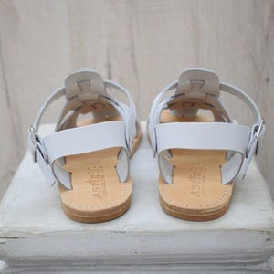 May include: A pair of white leather sandals with a strappy design. The sandals have a buckle closure and a light brown sole. The sandals are displayed on a white surface.
