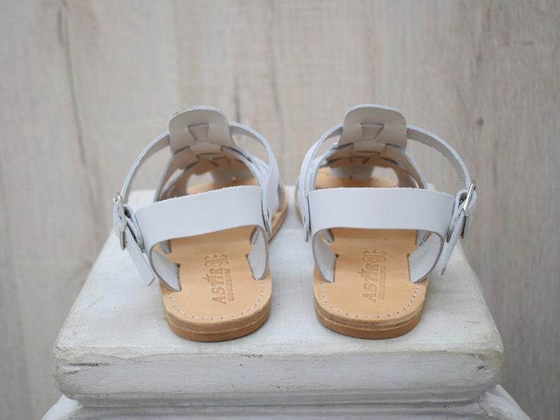 May include: A pair of white leather sandals with a strappy design. The sandals have a buckle closure and a light brown sole. The sandals are displayed on a white surface.