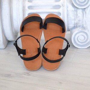 K&ouml;nnte beinhalten: Ein Paar schwarze Ledersandalen mit braunen Sohlen. Die Sandalen haben ein schlichtes Design mit einem breiten Riemen &uuml;ber dem Fu&szlig; und einem Kn&ouml;chelriemen mit einer silbernen Schnalle. Die Sohlen sind mit dem Oberteil vern&auml;ht.