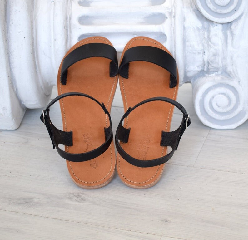 K&ouml;nnte beinhalten: Ein Paar schwarze Ledersandalen mit braunen Sohlen. Die Sandalen haben ein schlichtes Design mit einem breiten Riemen &uuml;ber dem Fu&szlig; und einem Kn&ouml;chelriemen mit einer silbernen Schnalle. Die Sohlen sind mit dem Oberteil vern&auml;ht.