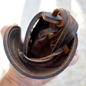Puede incluir: Primer plano de una sandalia de cuero marr&oacute;n, que muestra la suela, las costuras y los cordones de cuero. La sandalia tiene una suela marr&oacute;n oscuro con una superficie texturizada y un borde cosido. Los cordones son de cuero marr&oacute;n.
