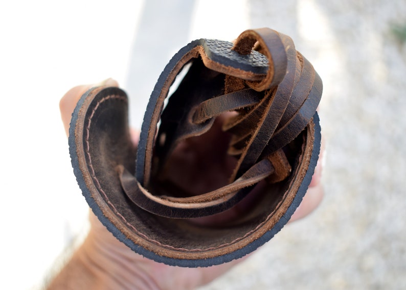 Puede incluir: Primer plano de una sandalia de cuero marr&oacute;n, que muestra la suela, las costuras y los cordones de cuero. La sandalia tiene una suela marr&oacute;n oscuro con una superficie texturizada y un borde cosido. Los cordones son de cuero marr&oacute;n.