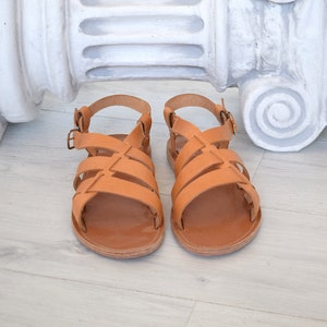 May include: A pair of tan leather sandals with multiple straps and adjustable buckles. The sandals have a flat sole and a classic design. The footwear is displayed on a light-colored wooden surface, with a white architectural element in the background.