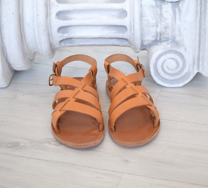 May include: A pair of tan leather sandals with multiple straps and adjustable buckles. The sandals have a flat sole and a classic design. The footwear is displayed on a light-colored wooden surface, with a white architectural element in the background.