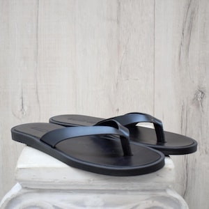May include: A pair of black leather flip-flops. The sandals feature a simple design with a flat sole and a thong strap. The flip-flops are displayed on a white surface against a light wood background.