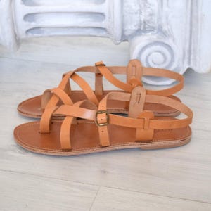 Op de afbeelding: Een paar bruinleren sandalen met meerdere bandjes en een gespsluiting. De sandalen hebben een platte zool en zijn geschikt voor warm weer. Ze worden getoond op een lichtgekleurd oppervlak.