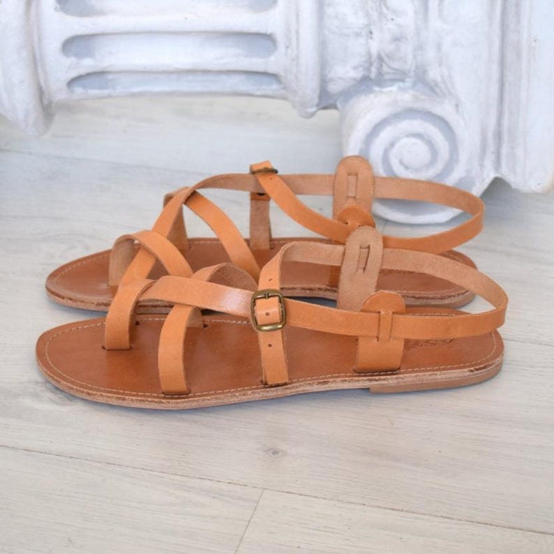Op de afbeelding: Een paar bruinleren sandalen met meerdere bandjes en een gespsluiting. De sandalen hebben een platte zool en zijn geschikt voor warm weer. Ze worden getoond op een lichtgekleurd oppervlak.