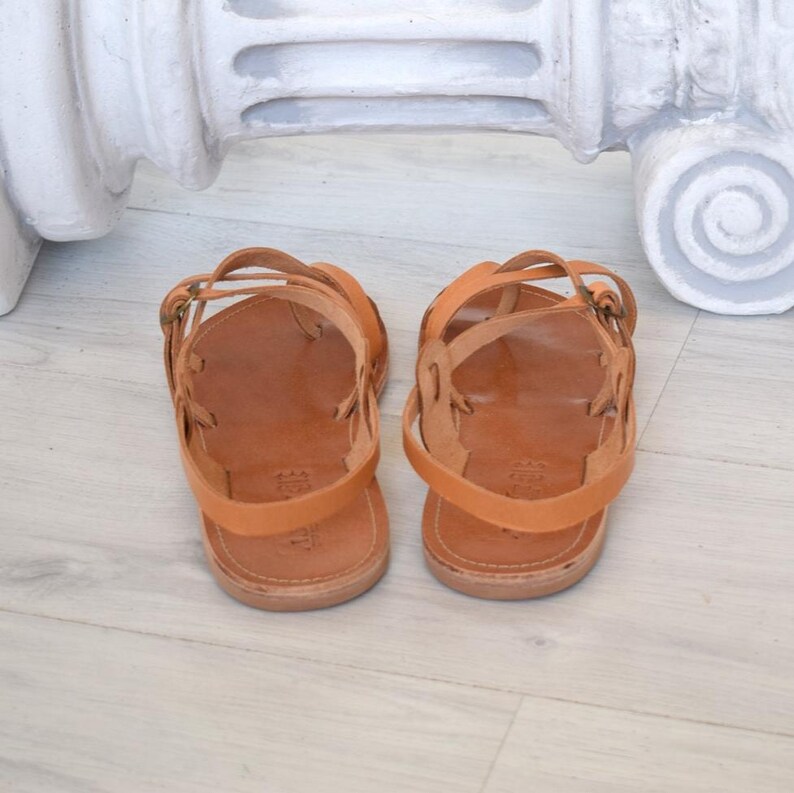 Op de afbeelding: Een paar lichtbruine leren sandalen met meerdere bandjes en gespen. De sandalen worden gepresenteerd op een lichtgekleurde houten vloer. Het ontwerp heeft een eenvoudige, klassieke stijl.