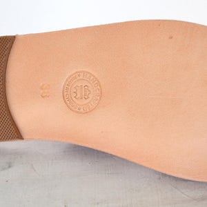 May include: A tan leather sandal sole featuring a brown textured heel pad and a circular embossed stamp in the center with the number 38. The smooth, light-colored surface of the sole shows natural grain and a rounded toe shape for footwear.