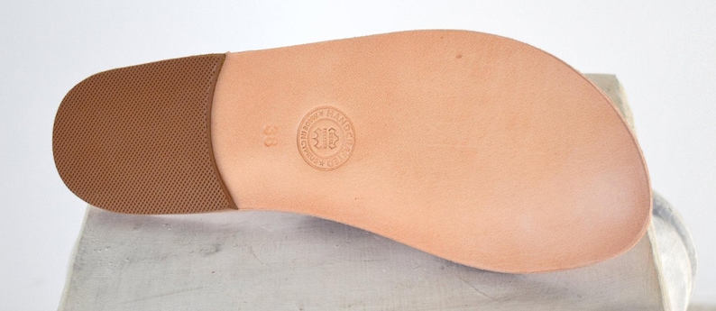 May include: A tan leather sandal sole featuring a brown textured heel pad and a circular embossed stamp in the center with the number 38. The smooth, light-colored surface of the sole shows natural grain and a rounded toe shape for footwear.