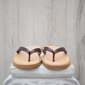 May include: A pair of brown leather flip-flops. The sandals have a light tan footbed, a dark brown thong strap, and a light brown sole. The flip-flops are displayed on a white surface against a light wood background.