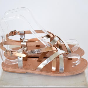 Può includere: Un paio di sandali in pelle color oro rosa e argento con cinturini incrociati. I sandali hanno una suola piatta e un cinturino alla caviglia con fibbia.