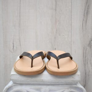 May include: A pair of leather flip-flops with dark brown straps and light brown soles. The sandals are displayed on a white surface against a light wood-grain background. The footwear is designed for casual wear.