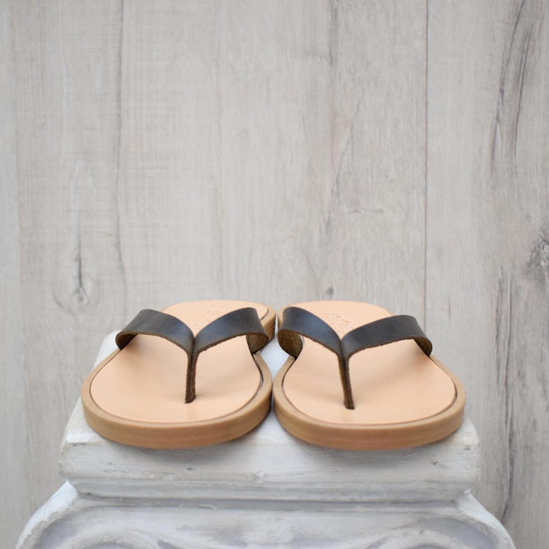 May include: A pair of leather flip-flops with dark brown straps and light brown soles. The sandals are displayed on a white surface against a light wood-grain background. The footwear is designed for casual wear.