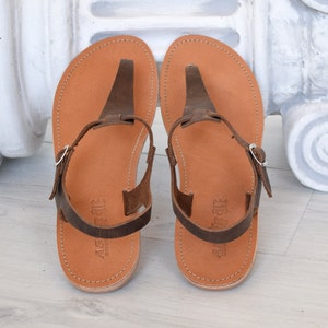 May include: A pair of brown leather sandals with a t-strap design. The sandals have a buckle fastening and are made from soft, supple leather.