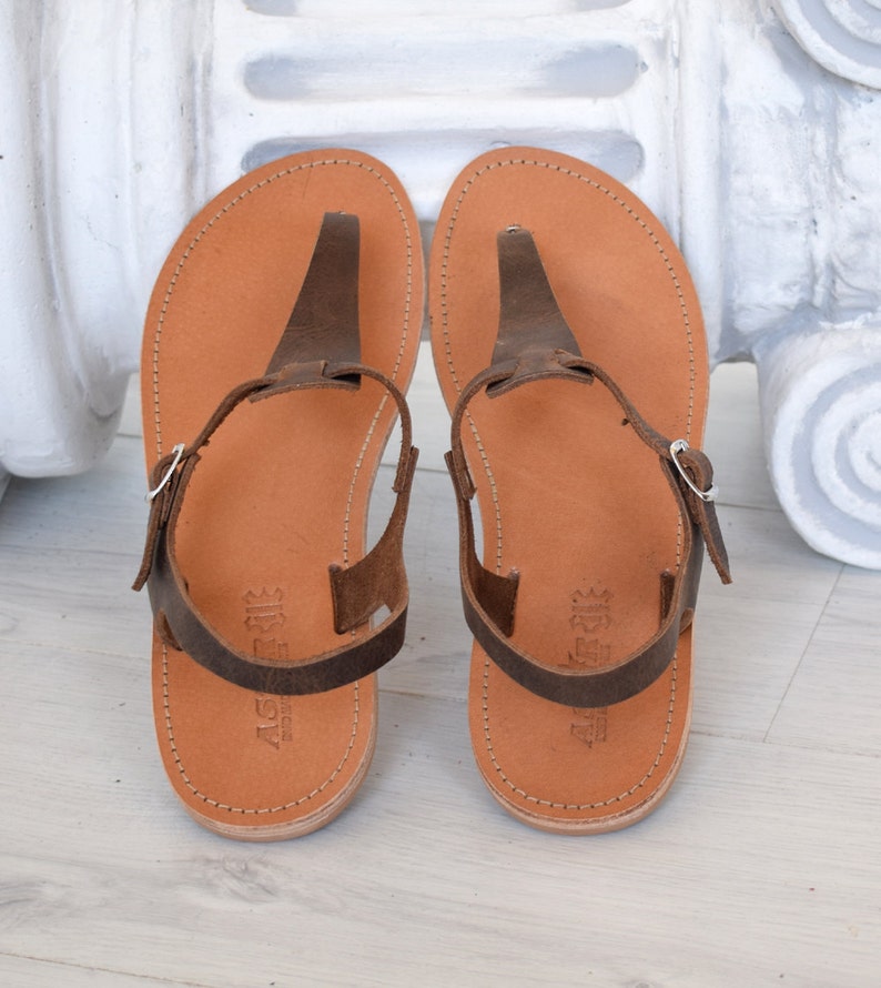 May include: A pair of brown leather sandals with a t-strap design. The sandals have a buckle fastening and are made from soft, supple leather.
