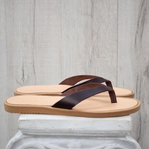 May include: A pair of brown leather flip-flops with a light tan sole. The straps are a darker shade of brown, and the sandals are displayed on a white pedestal against a light wood-grain background. The sandals are new.