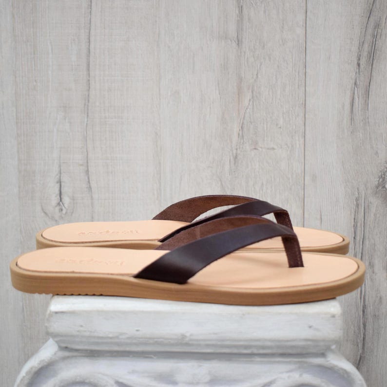 May include: A pair of brown leather flip-flops with a light tan sole. The straps are a darker shade of brown, and the sandals are displayed on a white pedestal against a light wood-grain background. The sandals are new.