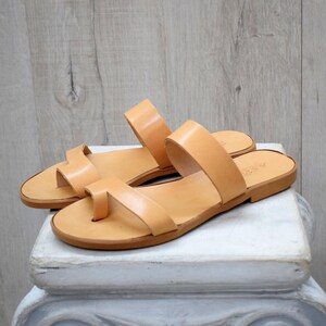 Könnte beinhalten: Ein Paar hellbraune Ledersandalen mit Zehenschlaufe und zwei Riemen. Die Sandalen werden auf einem weißen, dekorativen Sockel vor einem hellen Holzmaserungshintergrund präsentiert. Die Sandalen haben eine flache Sohle.