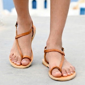 Peut inclure: Une paire de sandales en cuir beige au design minimaliste avec des brides fines, une boucle d'orteil et une petite fermeture à boucle métallique sur le côté. Les semelles plates sont fabriquées dans un matériau clair, offrant une chaussure simple.