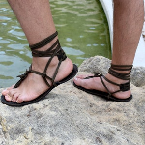 Puede incluir: Sandalias de cuero hechas a mano con correas marrones oscuras que se envuelven alrededor de los tobillos y los pies. Las sandalias tienen un dise&ntilde;o simple con una suela plana. La imagen muestra las sandalias en los pies de una persona, descansando sobre una roca cerca del agua.