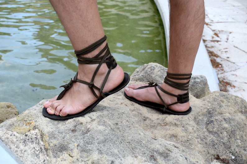 Puede incluir: Sandalias de cuero hechas a mano con correas marrones oscuras que se envuelven alrededor de los tobillos y los pies. Las sandalias tienen un dise&ntilde;o simple con una suela plana. La imagen muestra las sandalias en los pies de una persona, descansando sobre una roca cerca del agua.