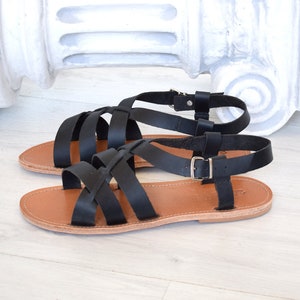 May include: A pair of black leather sandals with multiple straps and buckle closures. The sandals have a light brown sole and are displayed on a white wooden surface. The design is simple and elegant, suitable for warm weather.