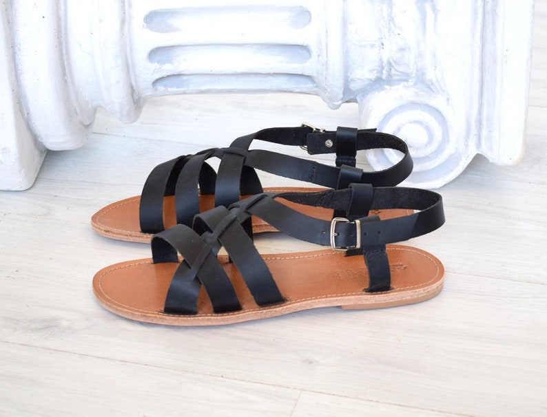 May include: A pair of black leather sandals with multiple straps and buckle closures. The sandals have a light brown sole and are displayed on a white wooden surface. The design is simple and elegant, suitable for warm weather.