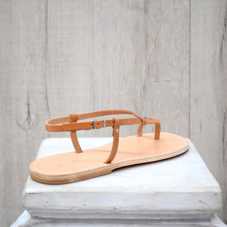 May include: A tan leather thong sandal featuring a flat sole, a toe post, and a thin adjustable ankle strap with a small silver-toned metal buckle. The sandal has visible white stitching along the edge of the sole and a smooth, light brown finish.