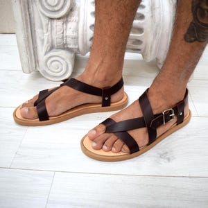 May include: A pair of dark brown leather sandals featuring a crisscross strap design over the top of the foot, a secure ankle strap with a silver-toned metal buckle, and a flat tan-coloured sole. The sandals are constructed with visible metal rivets.