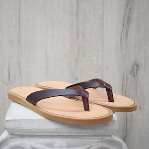 May include: A pair of brown leather flip-flops. They have a light tan footbed and a dark brown thong strap. The sandals have a light brown sole. They are displayed on a white surface against a light wood background.