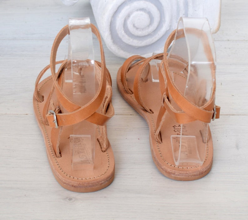Op de afbeelding: Een paar bruine leren sandalen met gekruiste banden en gespen. De sandalen liggen op een wit houten oppervlak.