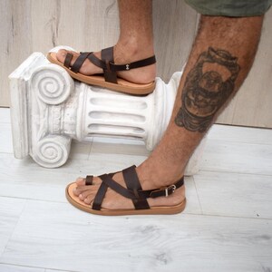 May include: Dark brown leather sandals featuring a toe loop design, multiple crisscrossing straps across the foot, and an adjustable ankle strap with a silver-toned metal buckle. The sandals have a flat, light tan sole and a simple construction.