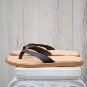 May include: A pair of black leather flip-flops with a light brown sole and straps. The sandals are displayed on a white surface against a light wood-grain background. The flip-flops are a neutral color and suitable for everyday wear.