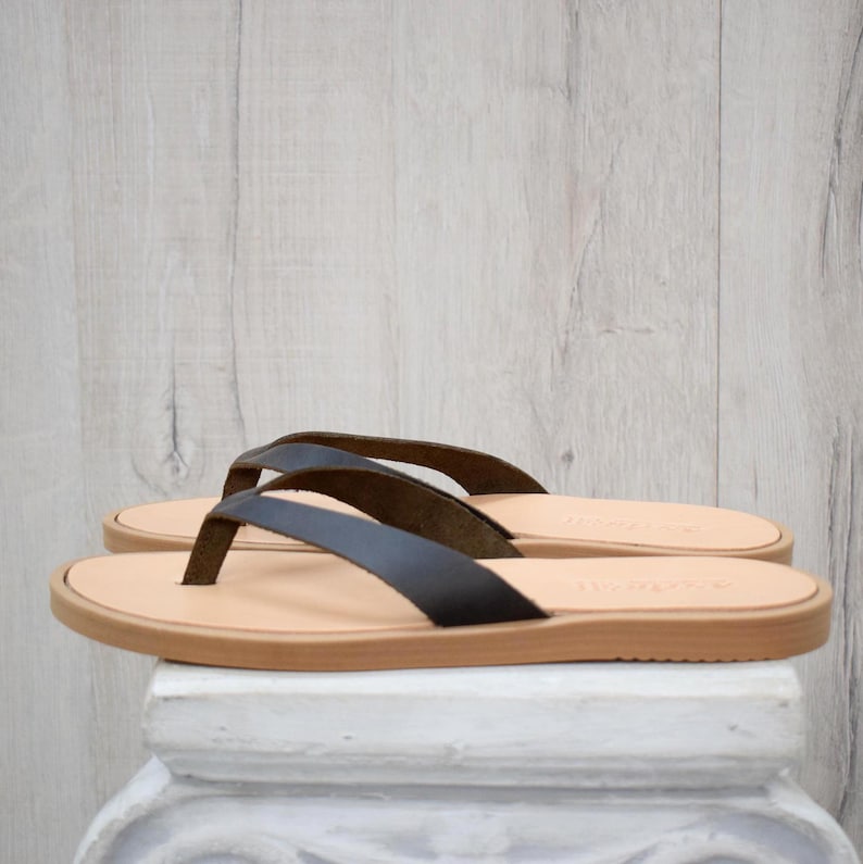 May include: A pair of black leather flip-flops with a light brown sole and straps. The sandals are displayed on a white surface against a light wood-grain background. The flip-flops are a neutral color and suitable for everyday wear.