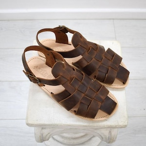 May include: A pair of brown leather sandals with woven straps and adjustable ankle straps. The sandals are displayed on a white surface. The leather has a distressed appearance, and the soles are a light tan color.