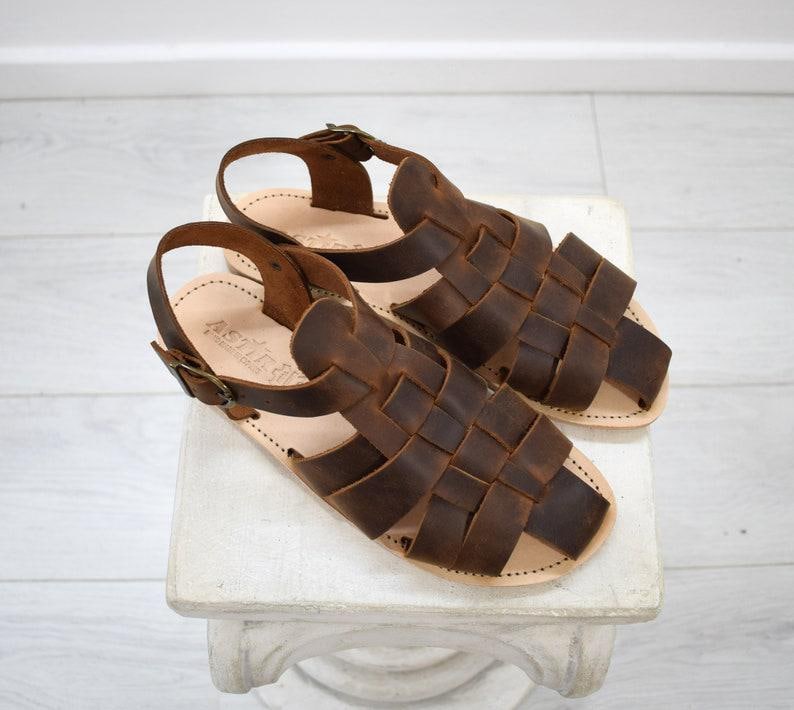 May include: A pair of brown leather sandals with woven straps and adjustable ankle straps. The sandals are displayed on a white surface. The leather has a distressed appearance, and the soles are a light tan color.