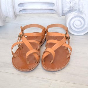Op de afbeelding: Een paar bruinleren sandalen met meerdere bandjes en een teensingel. De sandalen hebben een gespsluiting en worden gepresenteerd op een lichtgekleurd houten oppervlak. De sandalen zijn neutraal van kleur en lijken handgemaakt.