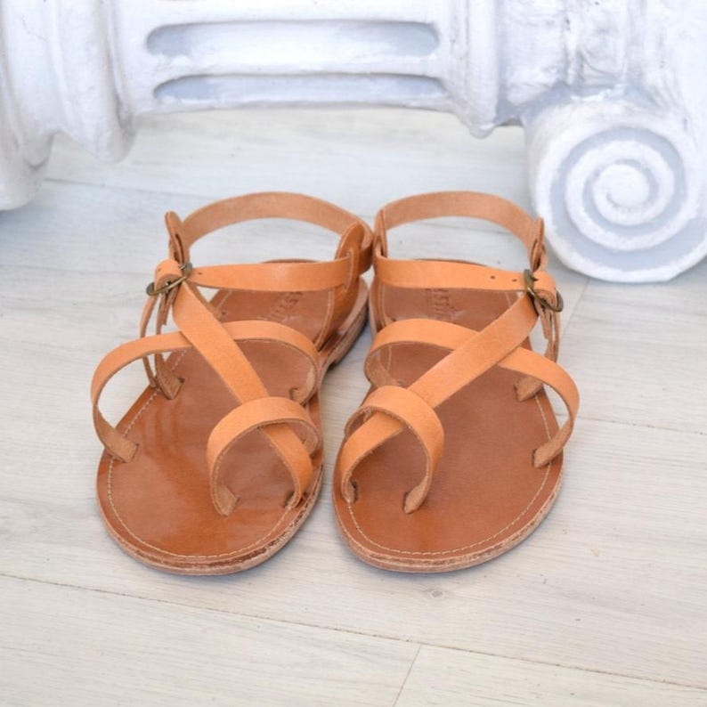 Op de afbeelding: Een paar bruinleren sandalen met meerdere bandjes en een teensingel. De sandalen hebben een gespsluiting en worden gepresenteerd op een lichtgekleurd houten oppervlak. De sandalen zijn neutraal van kleur en lijken handgemaakt.